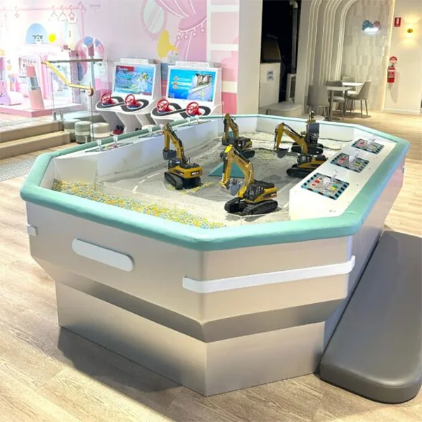 Interactive construction sandbox with toy excavators and control panels where children can operate miniature diggers in sand-filled play area at indoor family entertainment center