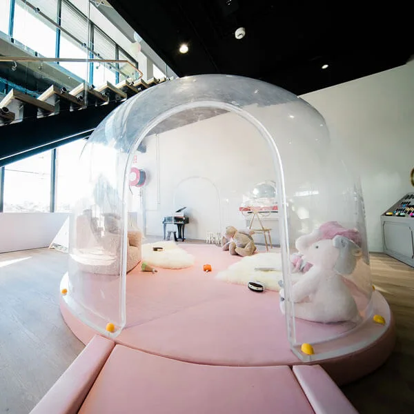 Clear inflatable bubble dome play area with pink padding, soft toys, and small table inside modern indoor facility with stairs visible
