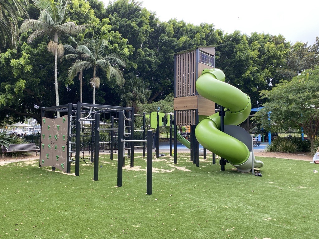 Renewed playground on synthetic grass with a timber tower, green spiral tunnel slide, climbing wall, monkey bars and rope frame, backed by tall palms and trees.