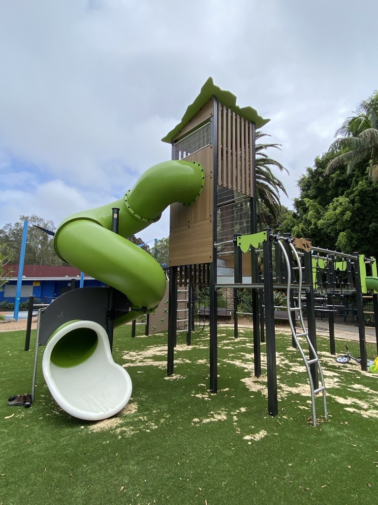 Tall timber climbing tower with a green spiral tunnel slide and a white open slide, monkey bars and a metal ladder over synthetic grass.