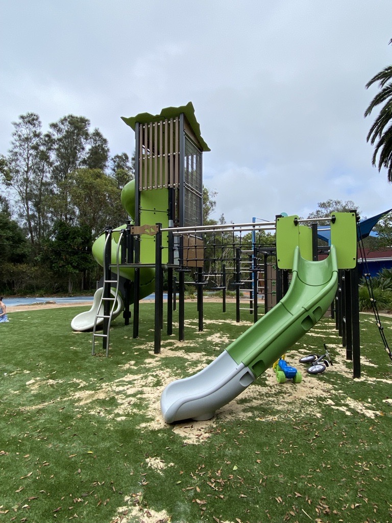 Side view of the multi-level play structure with a green and silver slide, monkey bars and elevated tower, with childrens ride-on toys on the grass.