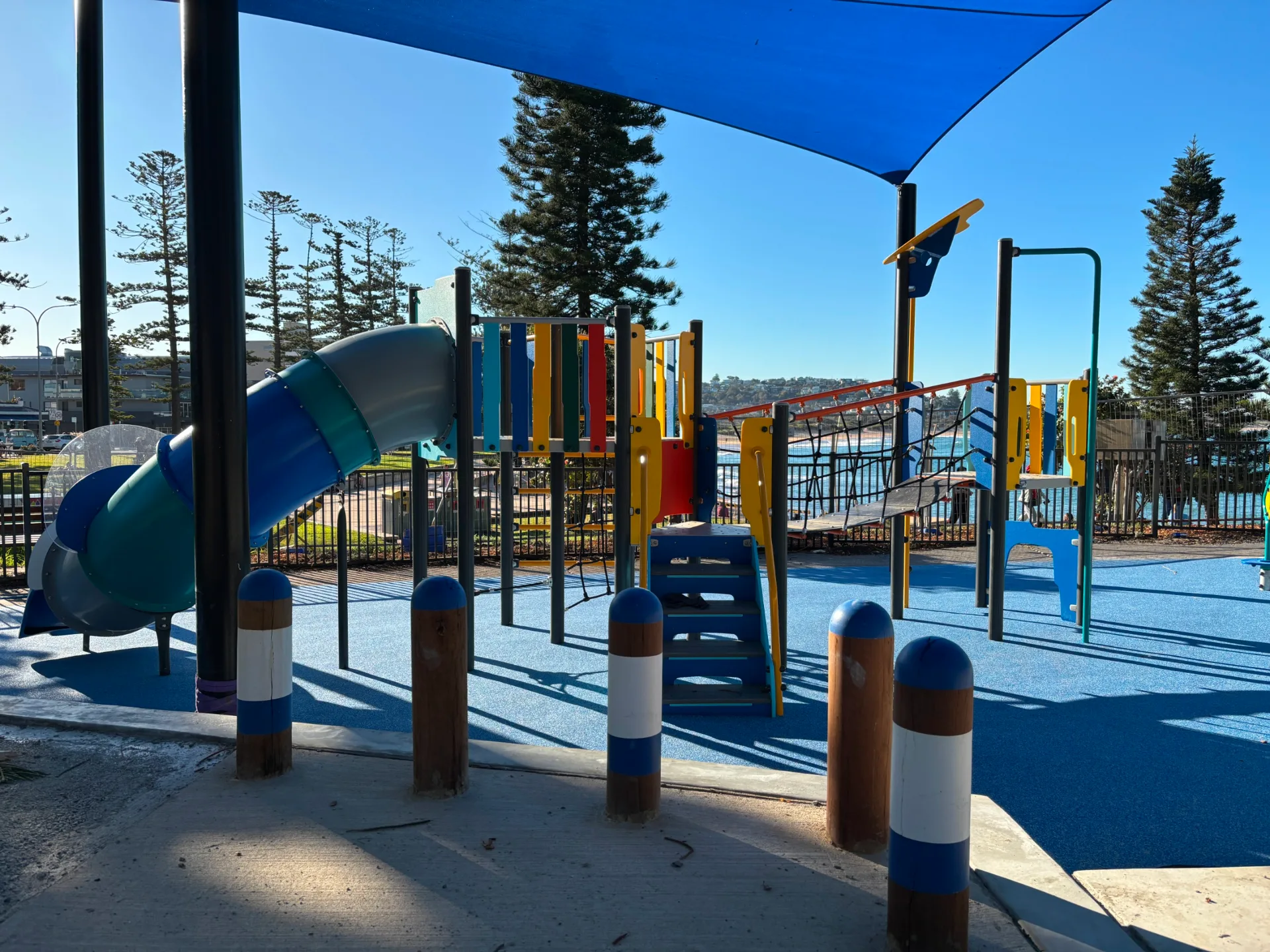 Colorful playground with blue slide, climbing structures, and shade sail over blue safety surfacing, surrounded by tall trees and safety bollards.