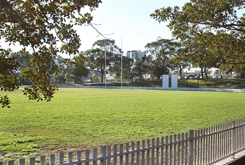 Open grassy sportsground at Camperdown Park with rugby goalposts, a timber picket fence in the foreground, mature trees and apartment buildings in the background.