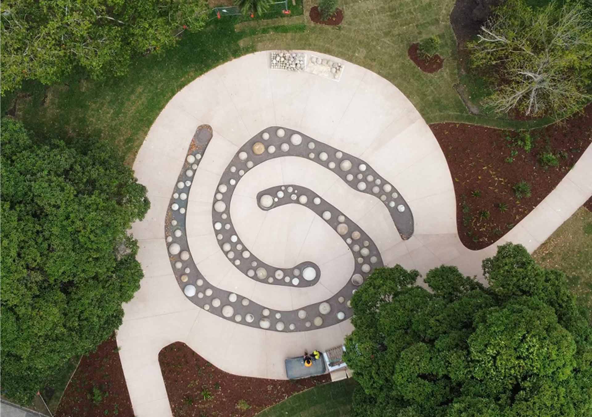 Aerial view of a paved circular plaza featuring a large spiral pattern of dark stepping stones, surrounded by garden beds and mature trees.