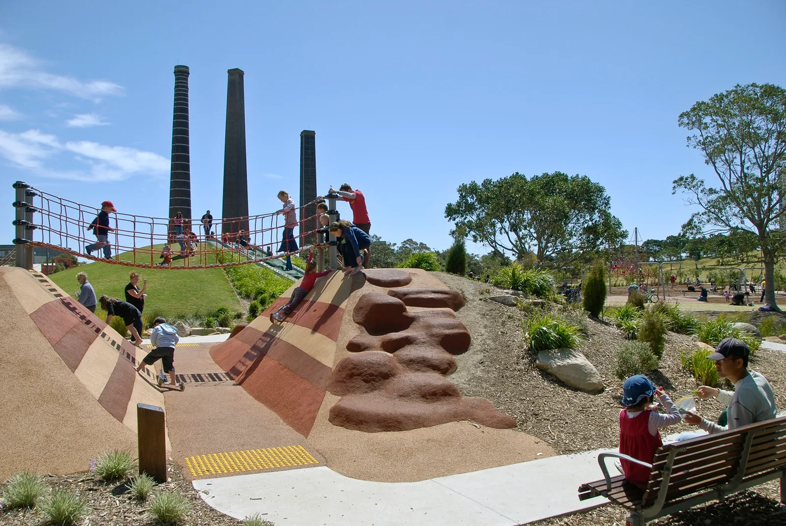 Modern adventure playground with sculptural climbing rocks, rope bridge, and grassy hill. Historic brick chimneys in background. Children playing on natural-toned equipment with landscaped surroundings.