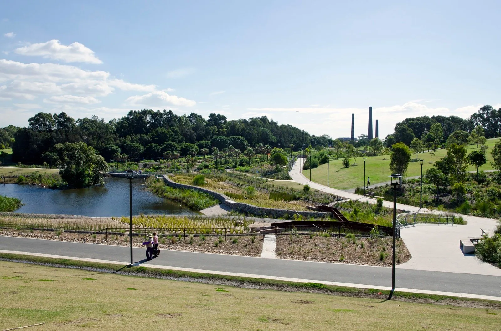 Landscaped park with winding pathways, a pond, grassy hills, and industrial chimneys in the distance under a partly cloudy sky.