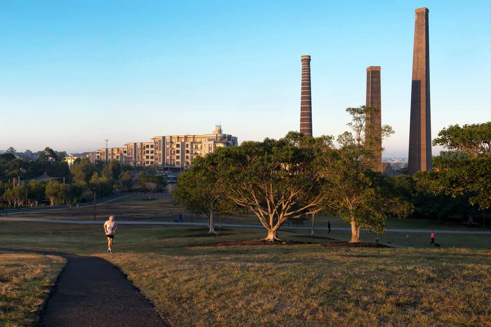 Open grassy park with walking paths and scattered trees, featuring historic brick chimneys in background and modern apartments, with jogger using paved trail at golden hour