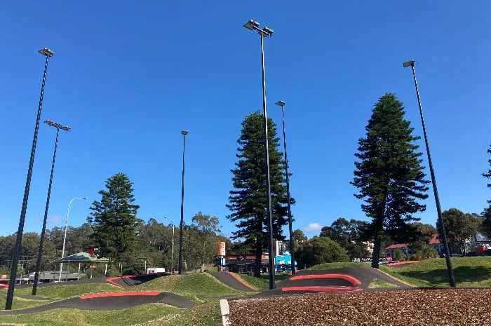 Modern pump track with curved red asphalt sections winding around grassy areas, surrounded by tall trees and lighting poles under a clear blue sky.