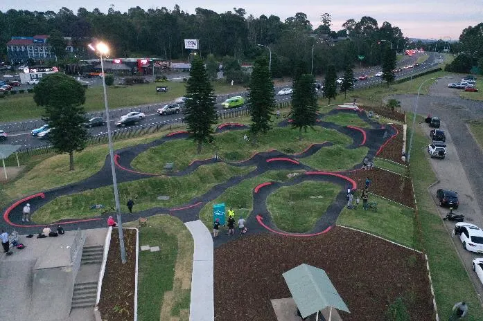 Aerial view of Leumeah Pump Track showing winding asphalt bike paths with red barriers through grassy terrain, surrounded by parking areas and trees at dusk