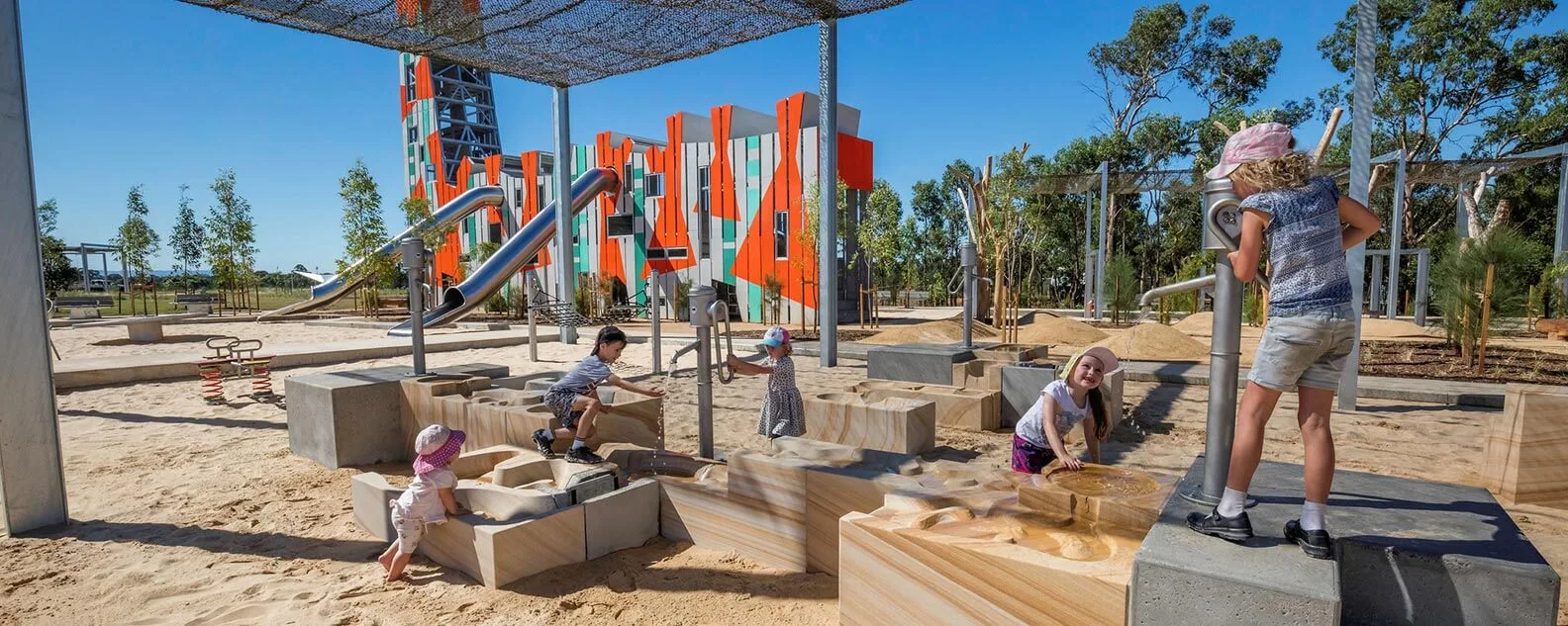 Children playing at a sand and water play station with silver hand pumps and sandstone channels. Shaded by a large overhead mesh.