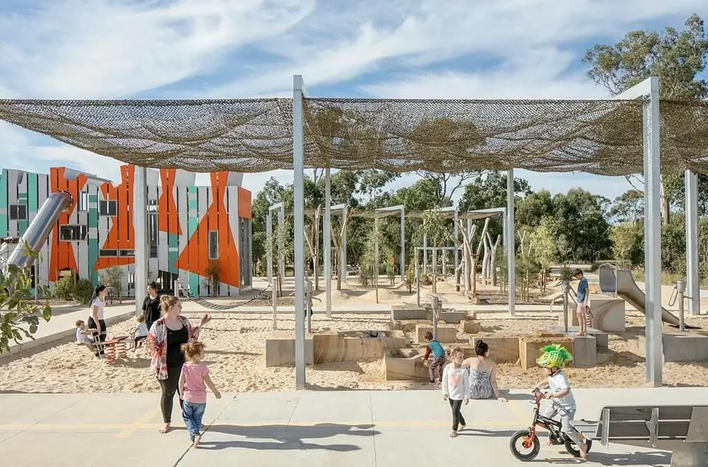 A large sand-pit playground featuring an expansive overhead shade sail, water play area, and children on bikes and scooters nearby.