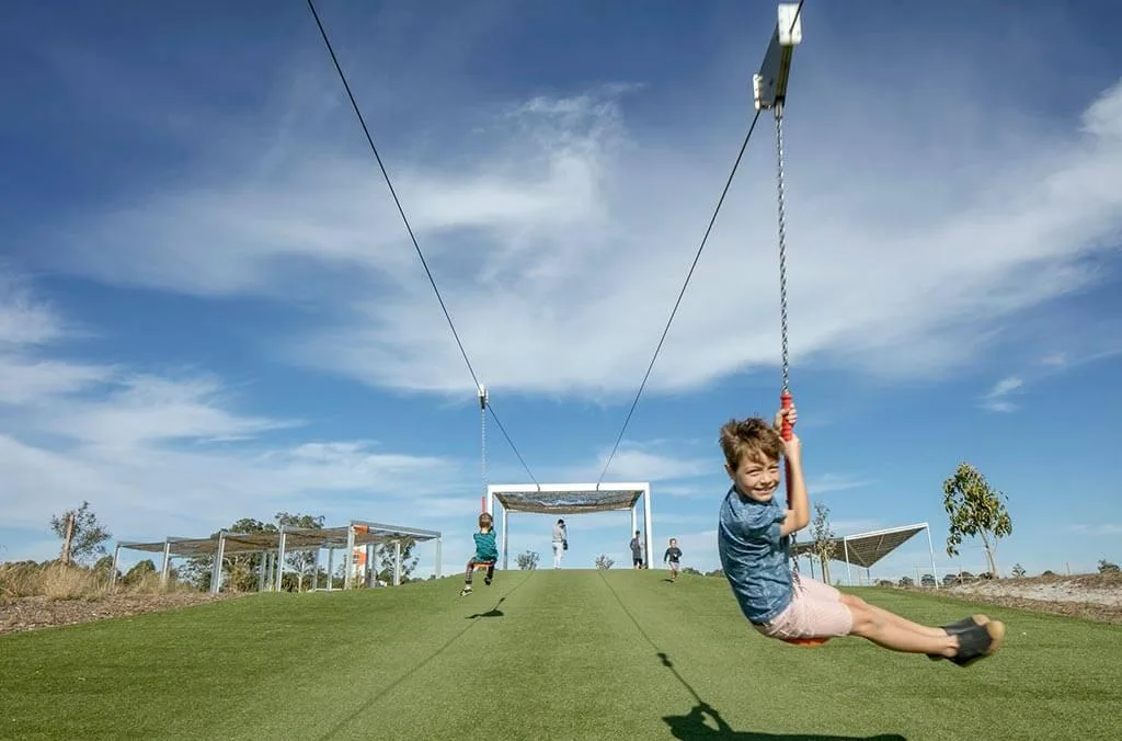 Two children riding long double zip lines over a green turf hill under a bright blue sky.