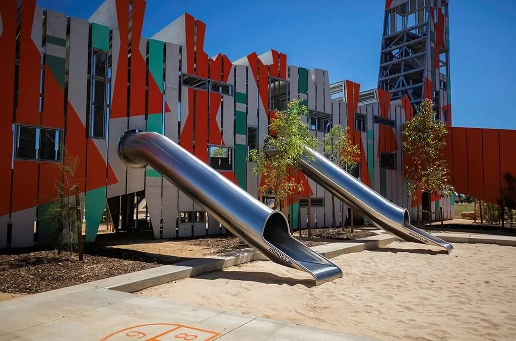 Close-up of two large stainless steel tunnel slides emerging from a vibrant orange and teal climbing structure over a sand base.