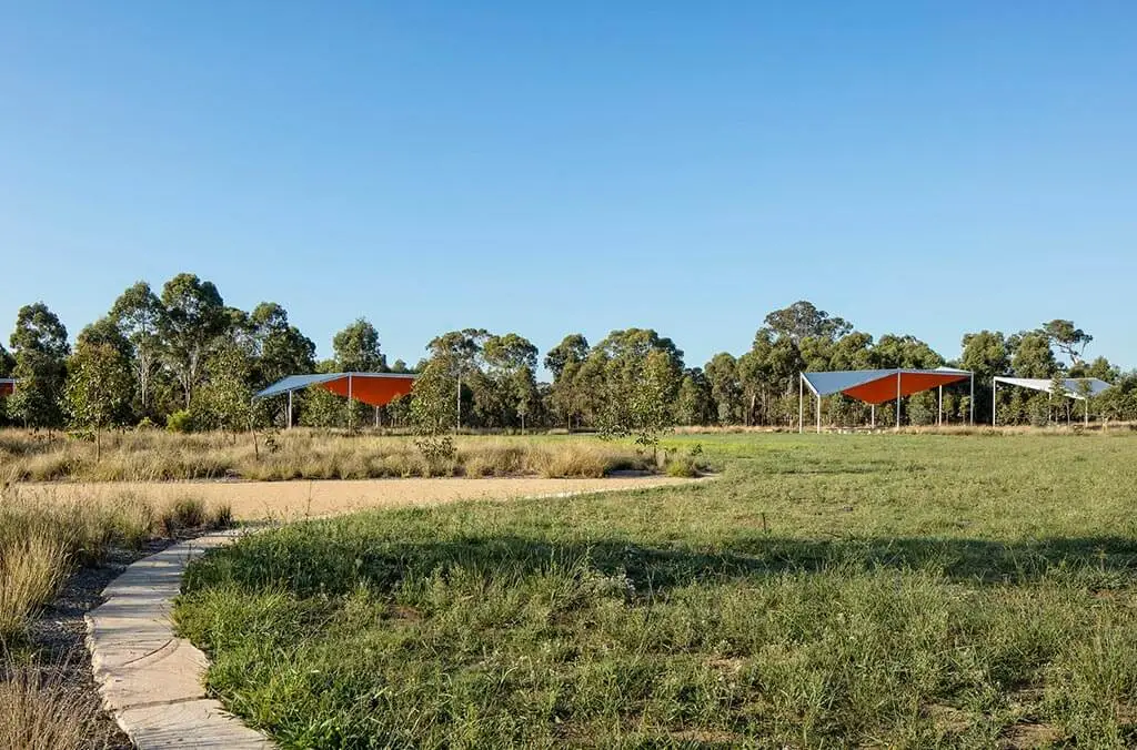 A winding stone path leading through a grassy field toward several large, modern orange shade pavilions and a line of trees.
