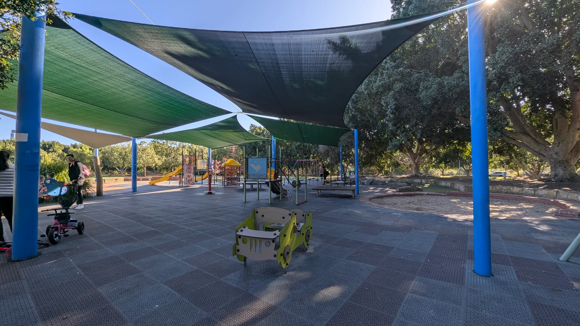 Shaded playground area with blue support posts and green shade sails covering rubber safety surfacing, featuring toddler play equipment and larger playground structures in background among mature trees.