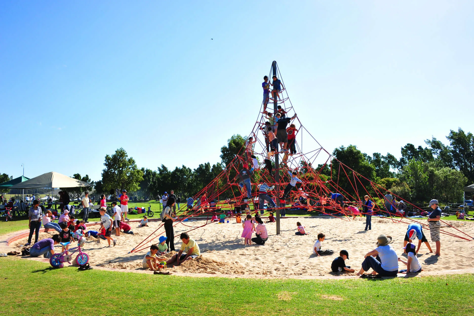 Large red rope climbing pyramid with children playing on multiple levels, surrounded by sandy play area with families gathered around on sunny day