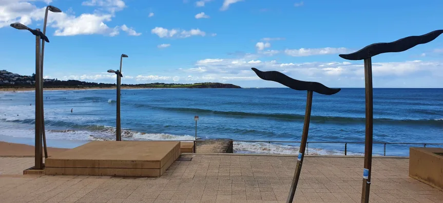 Dee Why Beach waterfront with modern shade structures, paved promenade, and calm blue waters perfect for family swimming and beach activities.