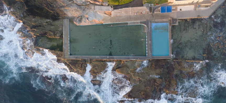 Aerial view of Dee Why ocean pool with green seawater, adjacent children's pool, and rocky coastline with crashing waves