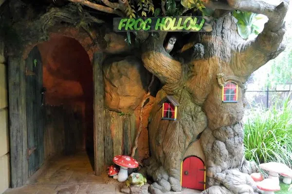 Whimsical tree-themed children's play area called "Frog Hollow" with carved wooden entrance, fairy doors, red mushroom decorations, and natural rock features creating an enchanted forest experience.