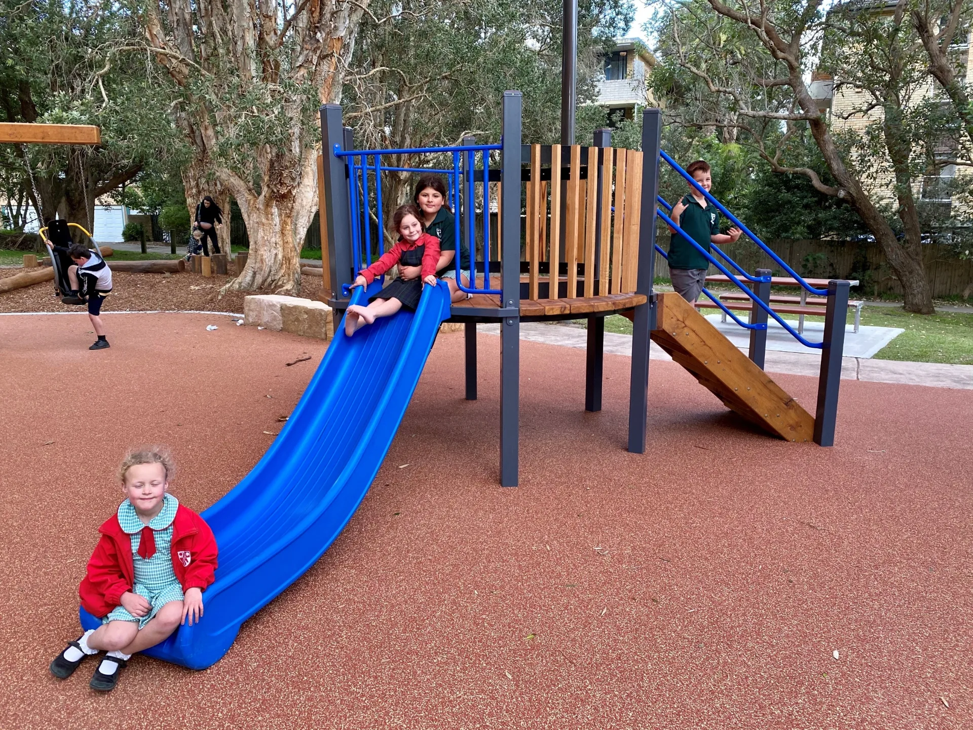 Children playing on a colorful playground with blue slide, wooden climbing structure, and monkey bars on rubber safety surfacing surrounded by mature trees