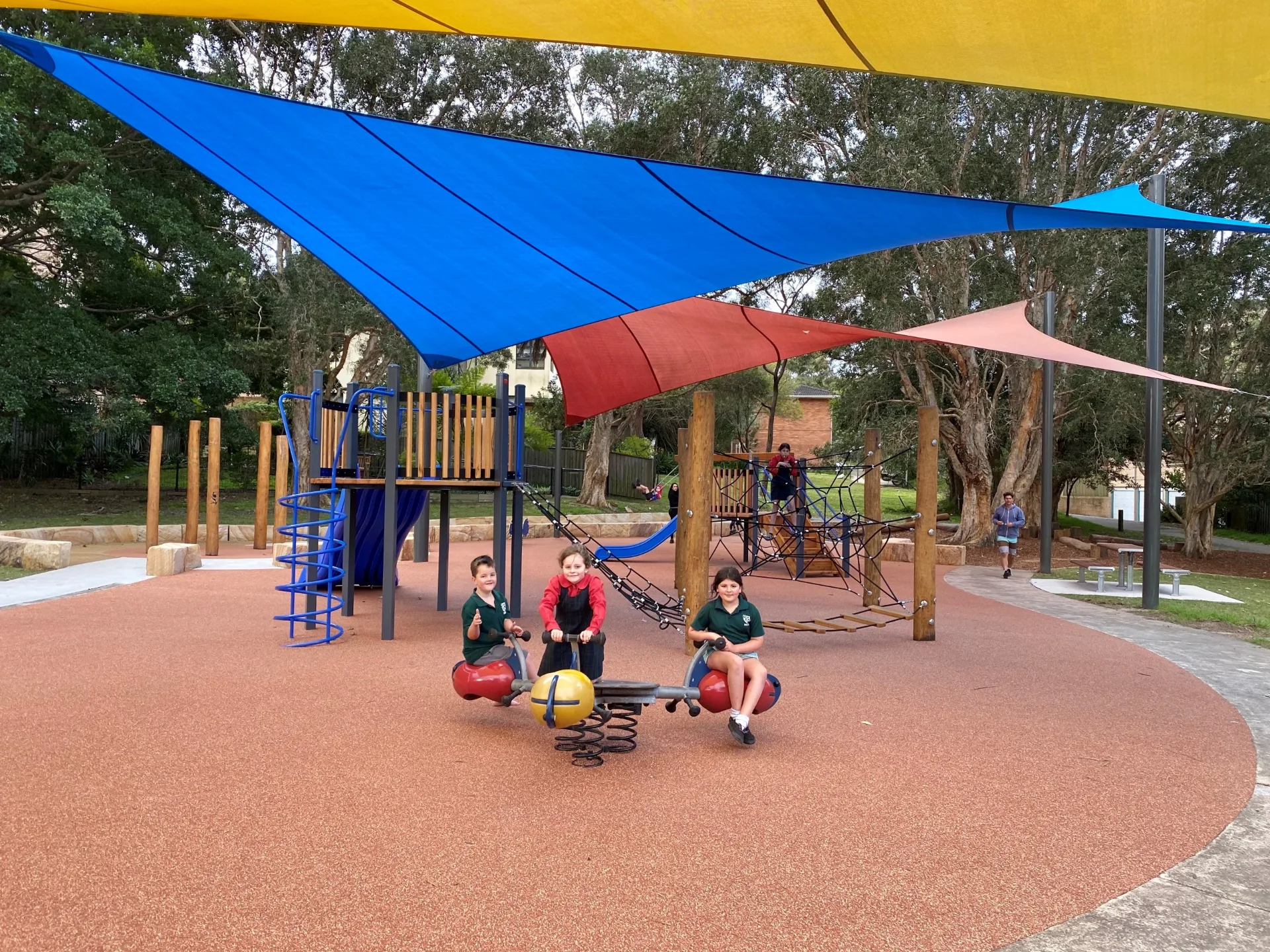 Modern playground with colorful shade sails, climbing equipment, slides, and spring riders on soft rubber surface surrounded by mature trees