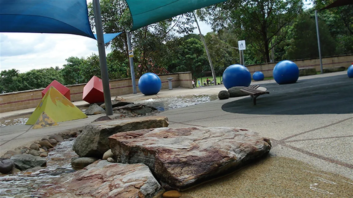 Water play area with large blue balls on platforms, rocky stream features, and colorful shade sails at Putney Park