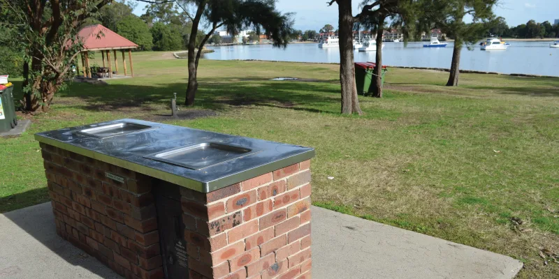 Brick barbecue station with metal cooking surface on concrete pad at waterfront park with boats visible on lake in background