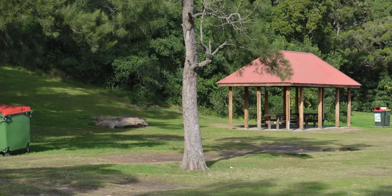 Spacious park with covered picnic pavilion featuring red roof and wooden posts, surrounded by green grass and mature trees providing natural shade