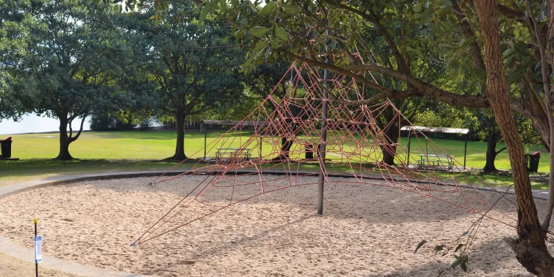 Large rope climbing pyramid structure on sand surface in tree-shaded park with open grassy fields and picnic areas in background