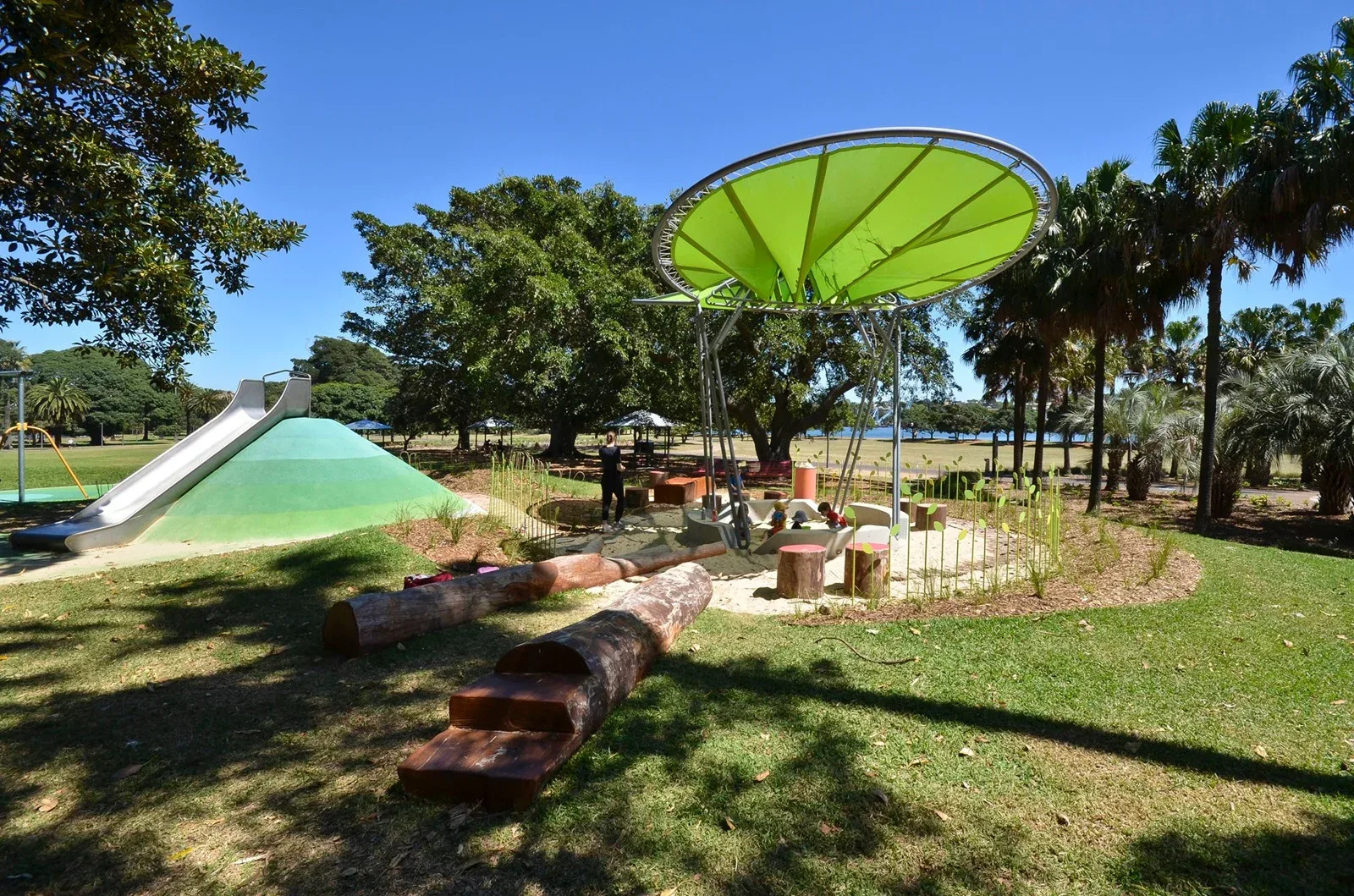 Modern playground with green fabric shade canopy over climbing structure, large green hill with slide, and fallen log balance beams on grassy field with palm trees in background.