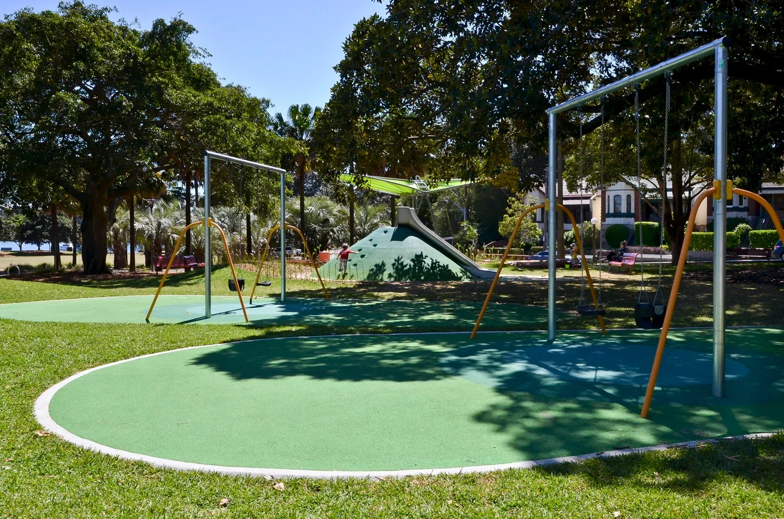 Modern playground with orange and silver swing sets on green rubber safety surfacing, featuring a large slide structure under mature shade trees in a sunny park setting.