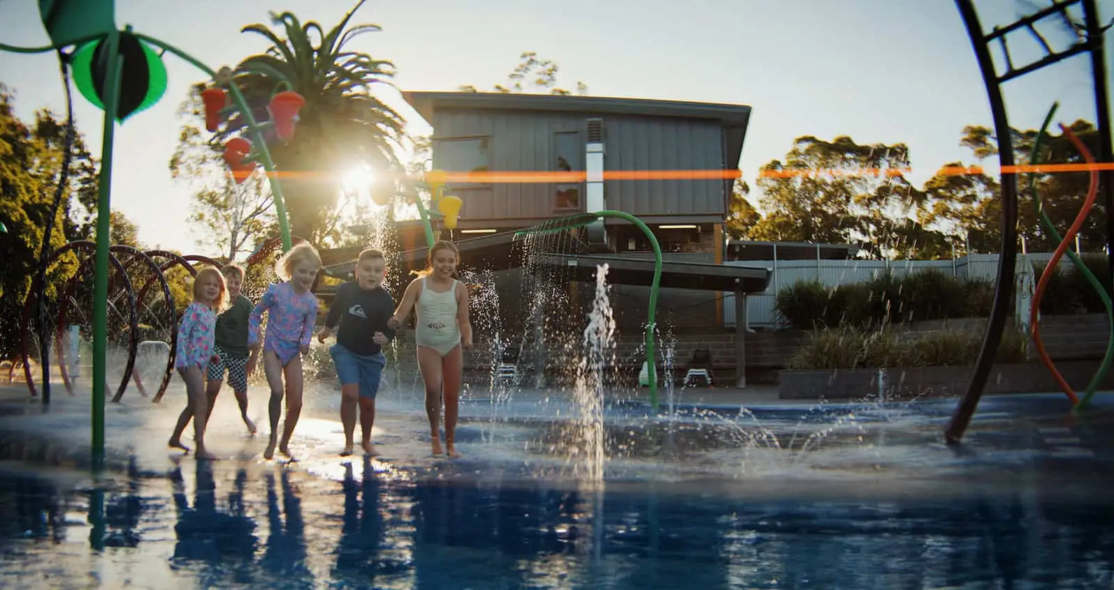 Children playing under the dump bucket and water jets at the on-site Splash Park, with soft non-slip surface and shade structures visible.