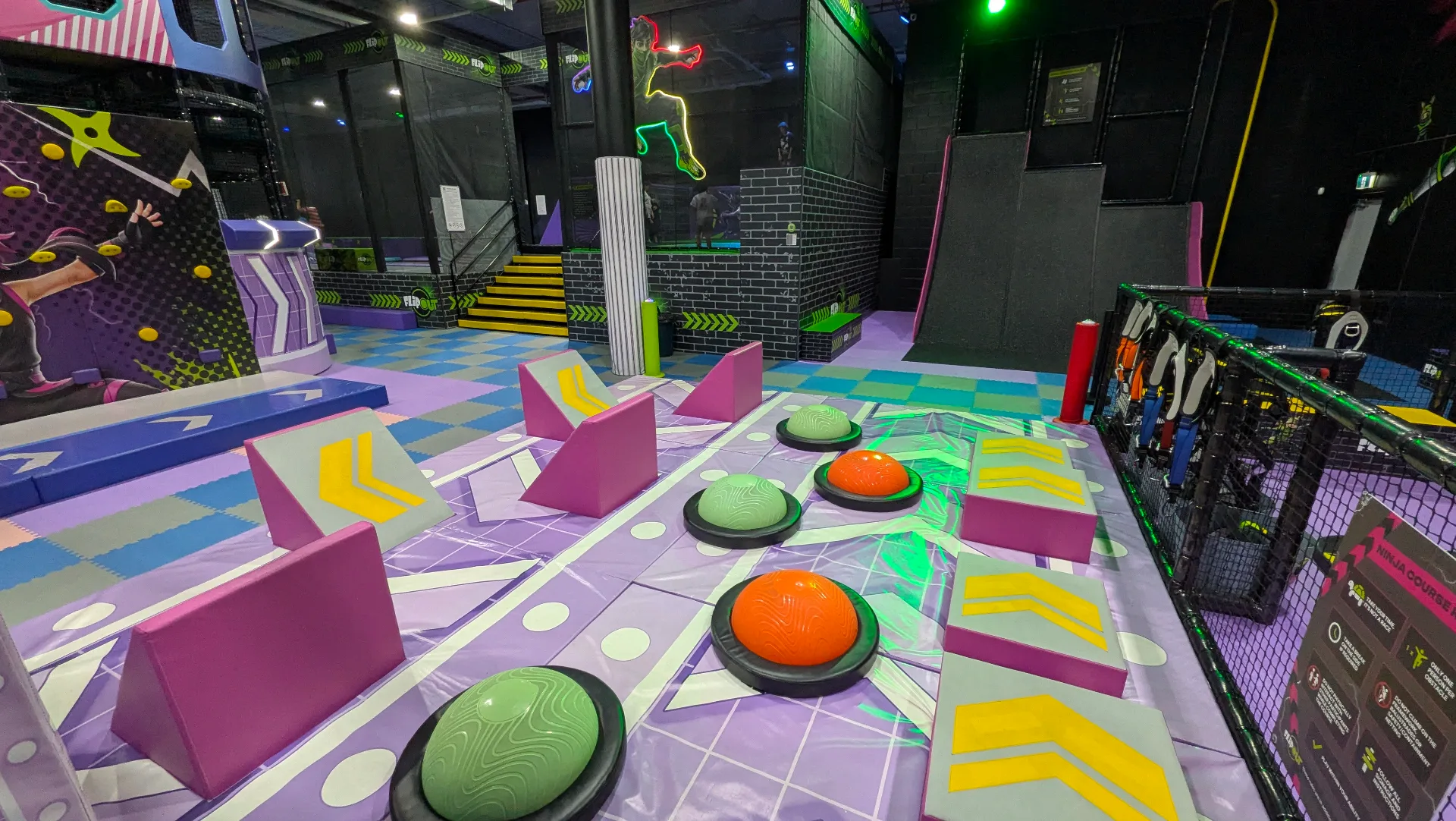 Indoor trampoline park with colorful foam obstacle course featuring large green and orange foam buttons, pink geometric blocks, and vibrant floor graphics in a neon-lit arena setting.