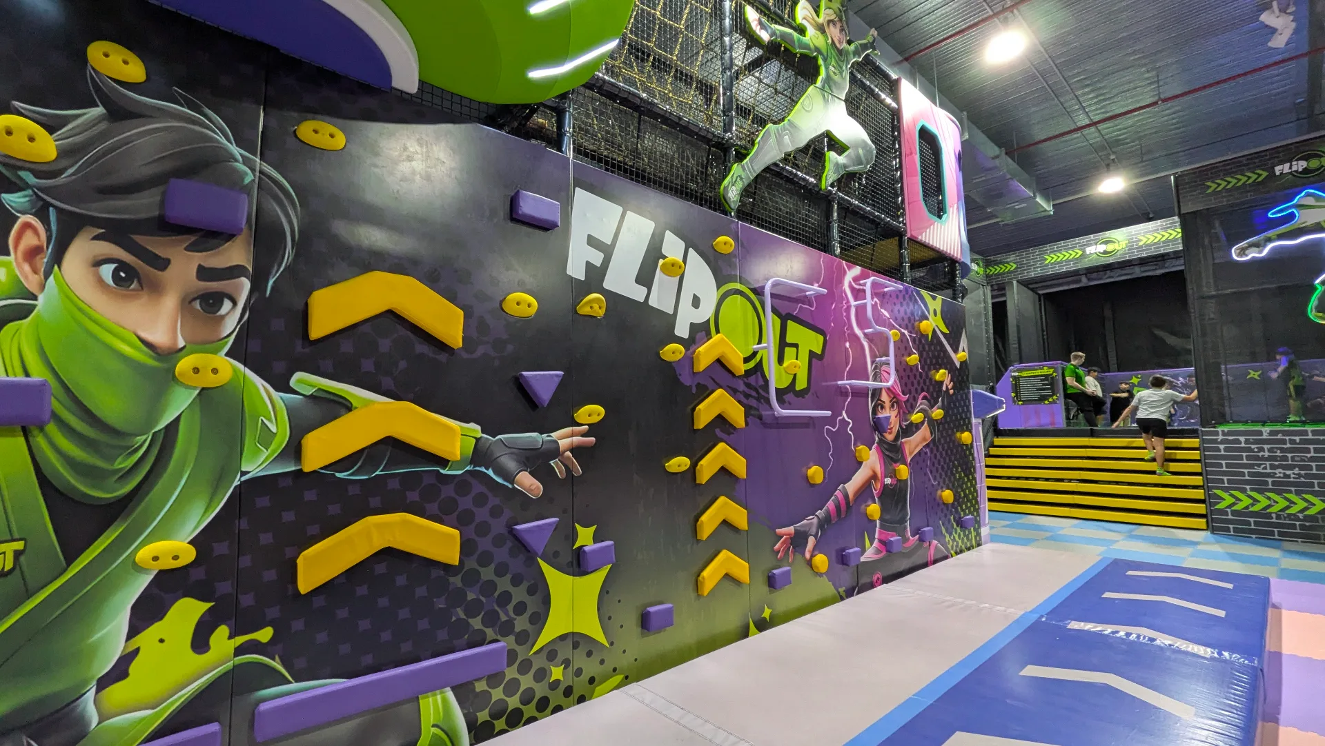 Indoor trampoline park with colorful ninja-themed wall mural featuring climbing holds, protective netting above, and blue safety mats on the floor.
