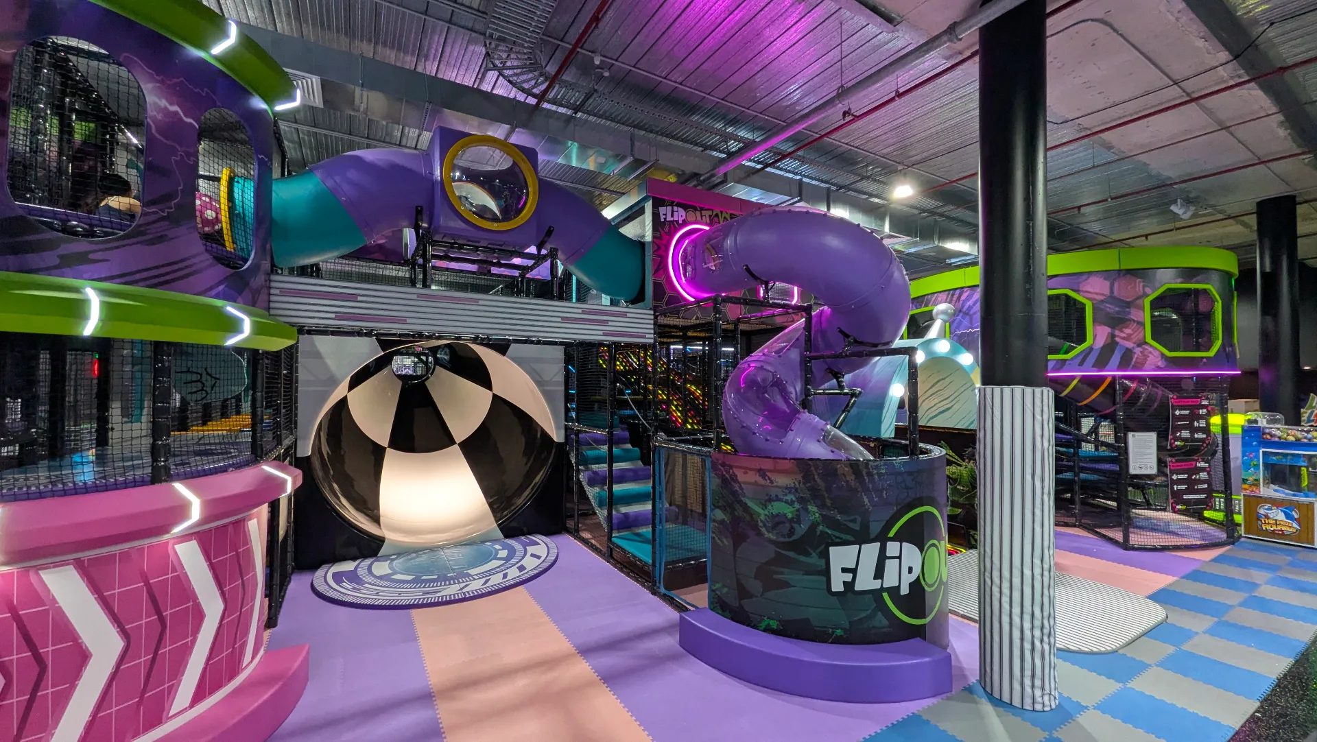 Indoor trampoline park with futuristic space theme featuring purple tube slides, multi-level climbing structures, and colorful padded safety surfaces throughout the play area.
