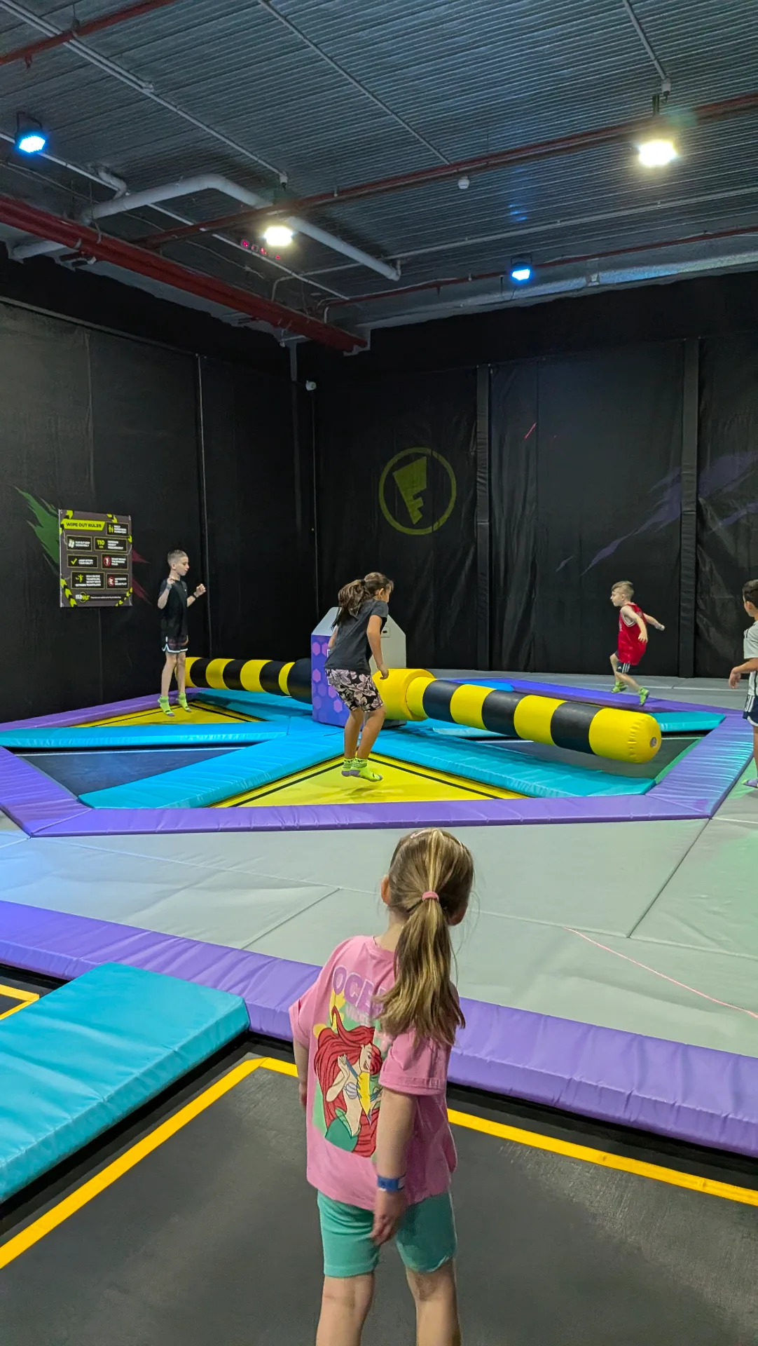 Indoor trampoline park with children playing on colorful trampolines and inflatable obstacle course, featuring yellow and black padded equipment in a vibrant recreational facility
