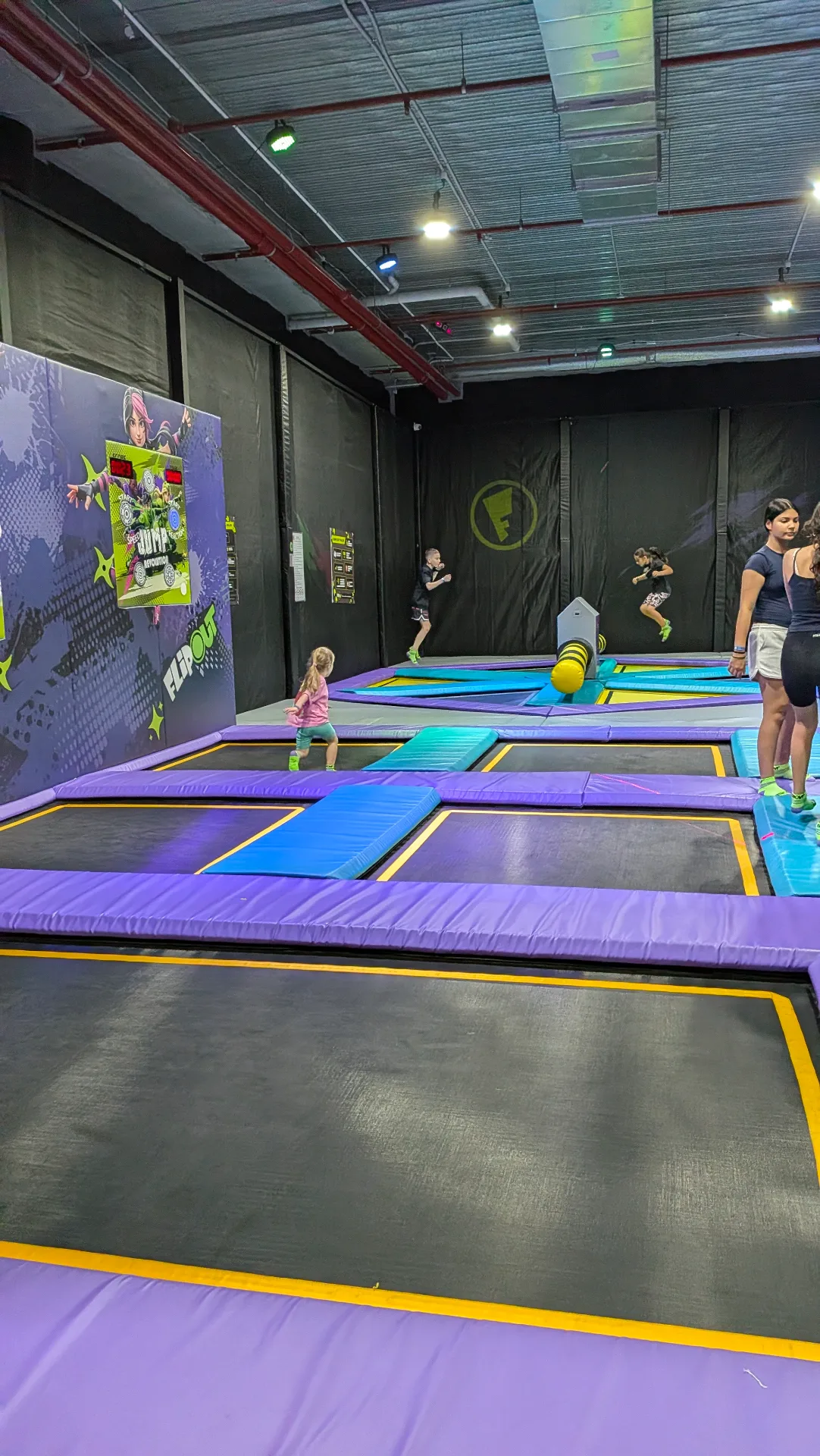 Indoor trampoline park with interconnected purple and blue trampolines, safety padding, and colorful wall graphics. Children and adults bouncing in a spacious facility with industrial ceiling.