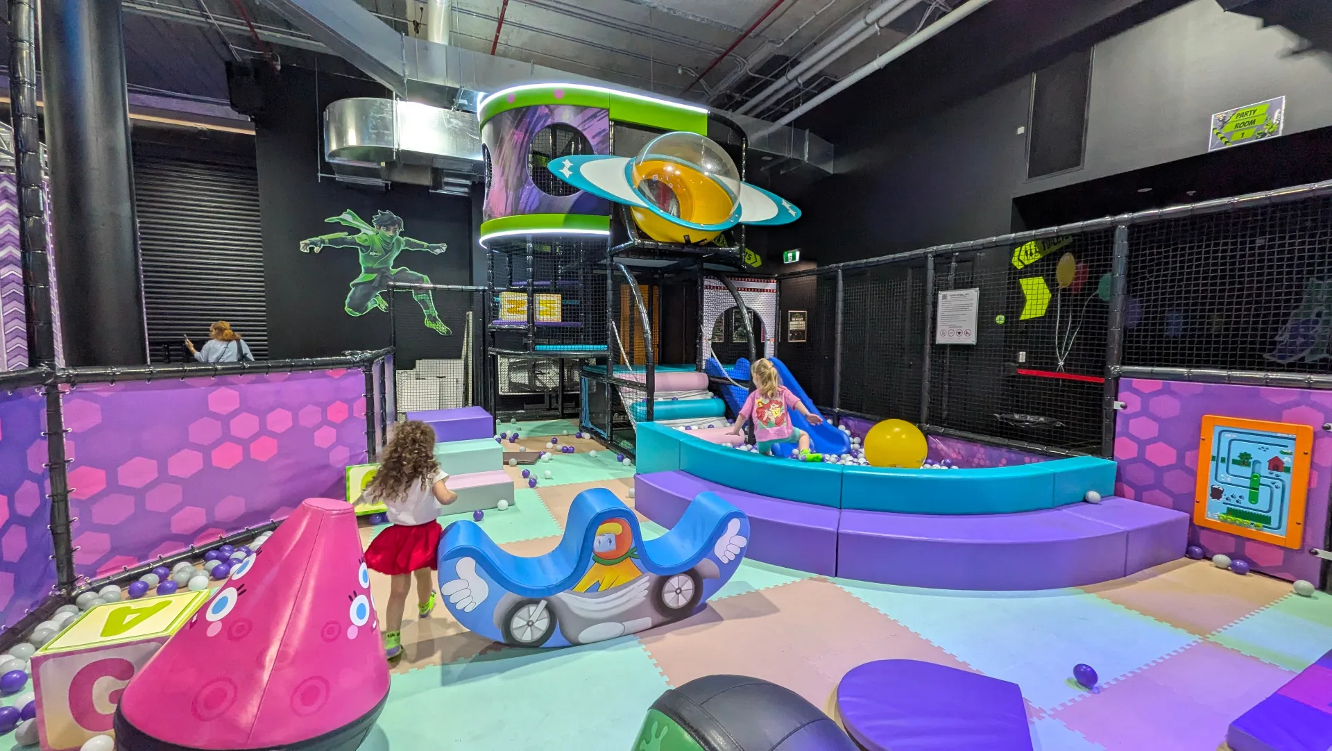 Colorful indoor toddler play area with soft foam climbing structures, ball pit, space-themed decorations, and padded flooring in a secure netted environment.