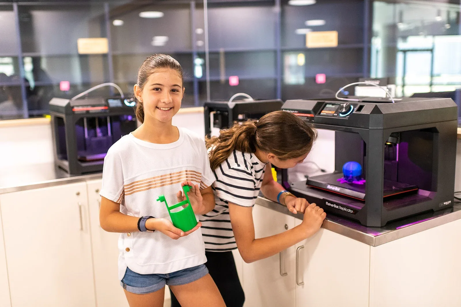 Two young girls interacting with 3D printers on a counter inside a modern makerspace, one proudly holding up a green plastic 3D-printed object.