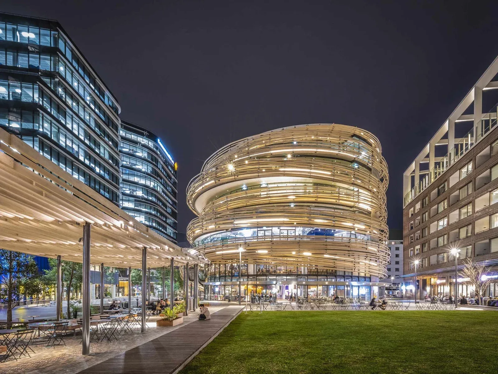 Vibrant, multi-story circular building wrapped in spiral wooden ribbons, illuminated at night next to a flat public green lawn with outdoor seating.