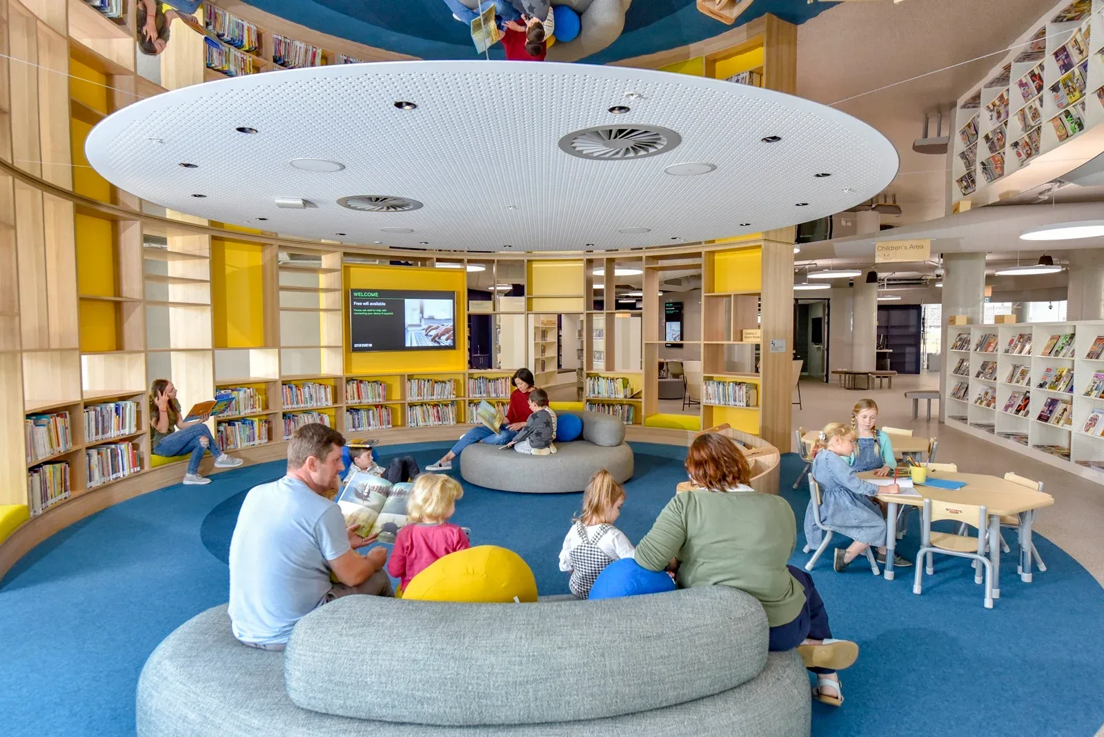 Bright, spacious children's library section with low curved shelving, soft blue carpeting, circular seating, and child-sized activity tables.