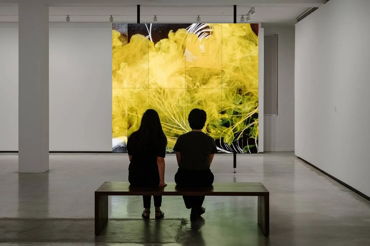 Bright, smoothly paved gallery room offering accessible paths, where two visitors sit on a wooden bench watching a large yellow digital projection.