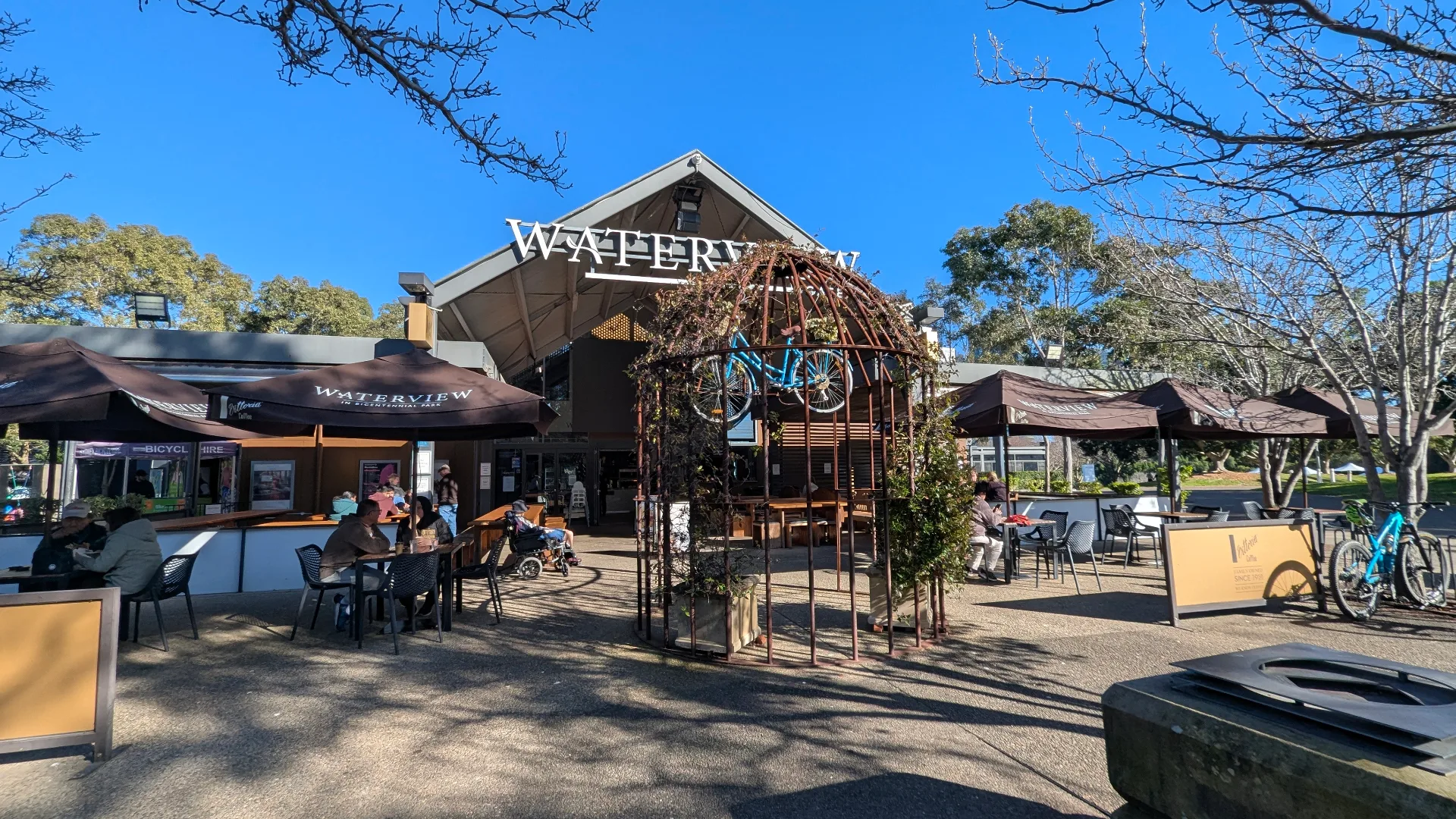 Waterview cafe with outdoor seating under umbrellas, decorative bicycle displays, and bicycle hire station surrounded by leafy trees on a sunny day.