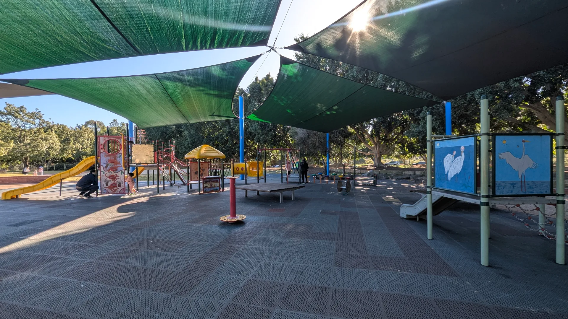 Shaded playground with colorful climbing equipment, slides, and educational panels featuring bird illustrations on rubberized surface surrounded by trees.