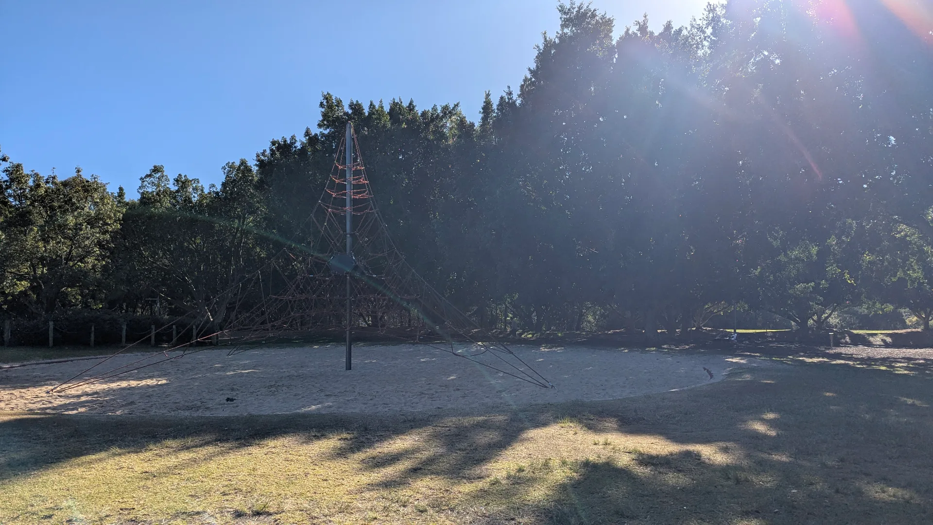 Large rope climbing pyramid on sandy playground surface with dense trees providing natural shade and yellow caution tape marking area boundaries.