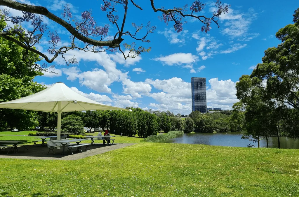 Sun-drenched park with shaded picnic shelter, manicured lawn, and serene lake views with city skyline backdrop at Bicentennial Park.