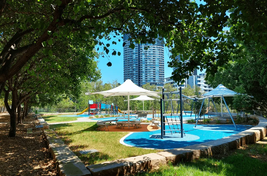 Shaded playground with blue splash pad, climbing equipment, and picnic shelter surrounded by mature trees, with high-rise buildings visible in background.