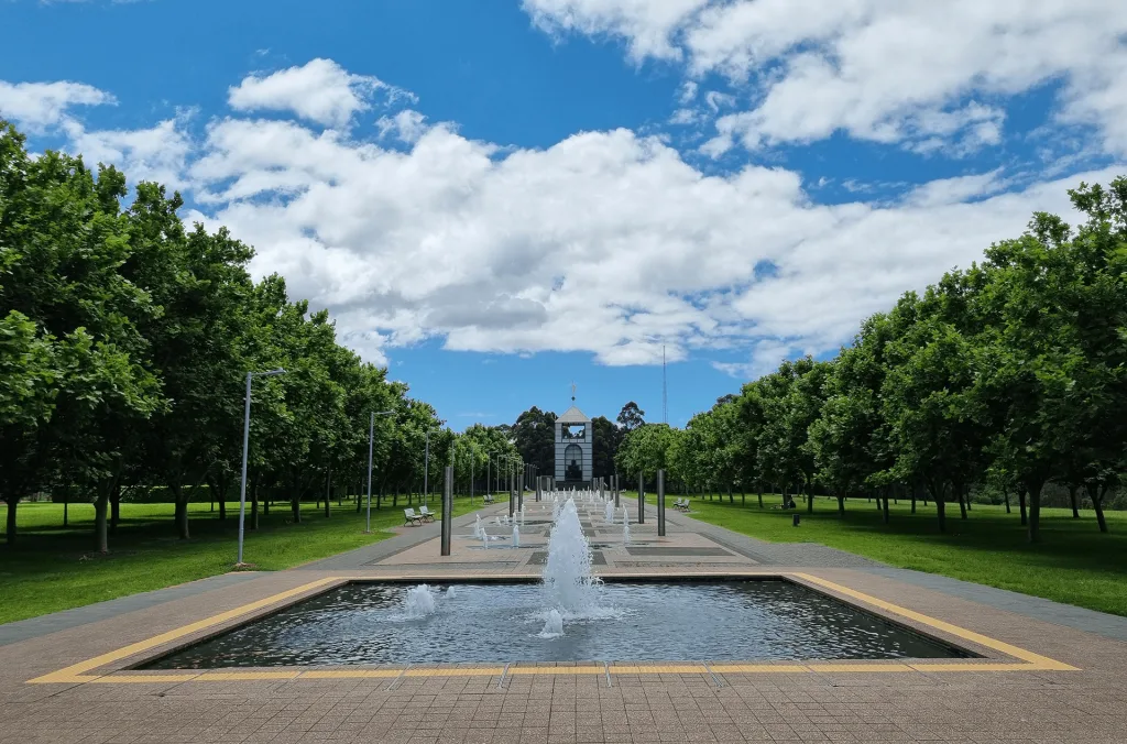 Tree-lined walkway with fountains leading to a historic building at Bicentennial Park, featuring paved paths suitable for strollers and wheelchairs.