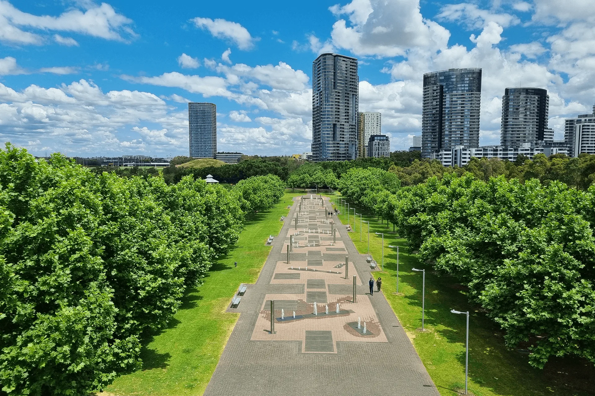 Aerial view of Bicentennial Park showing a long memorial pathway with geometric stone sections flanked by mature trees, leading toward modern high-rise buildings in the background.