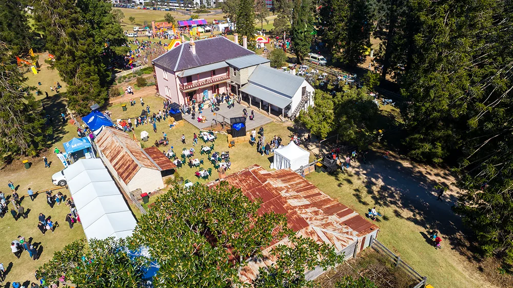 Aerial view of Bella Vista Farm showing a historic homestead with red-roofed outbuildings surrounded by families enjoying festival activities on expansive green lawns under mature shade trees.